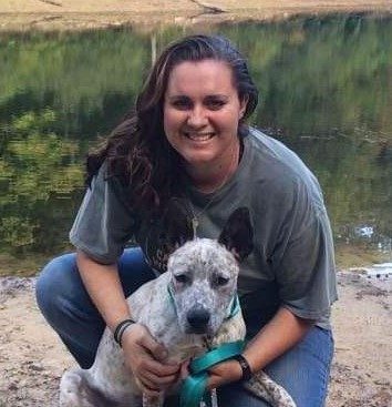 Stephanie Moss kneeling beside a speckled heeler at the edge of a lake